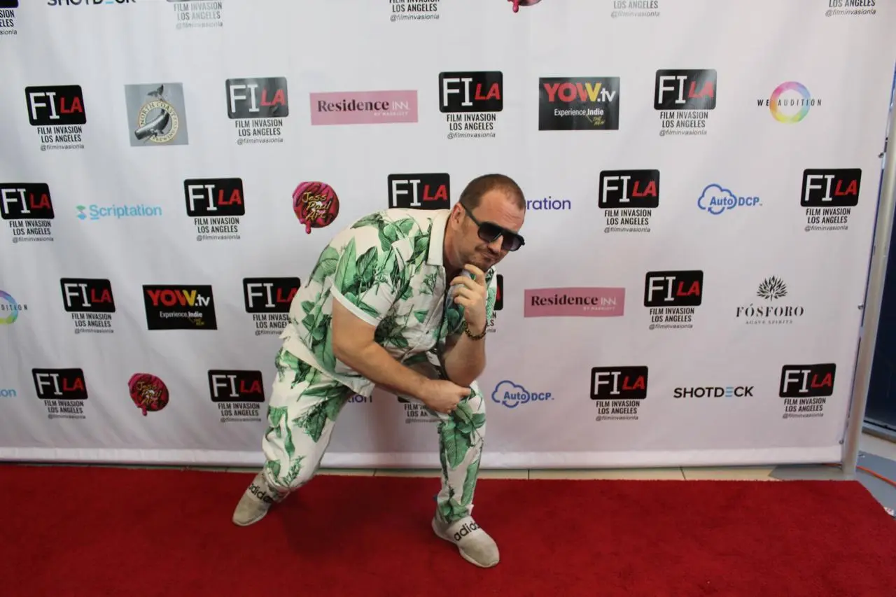 Matt Moeller in a green and white leaf-print suit and sunglasses, posing with a microphone on the red carpet in front of the Film Invasion Los Angeles step-and-repeat.
